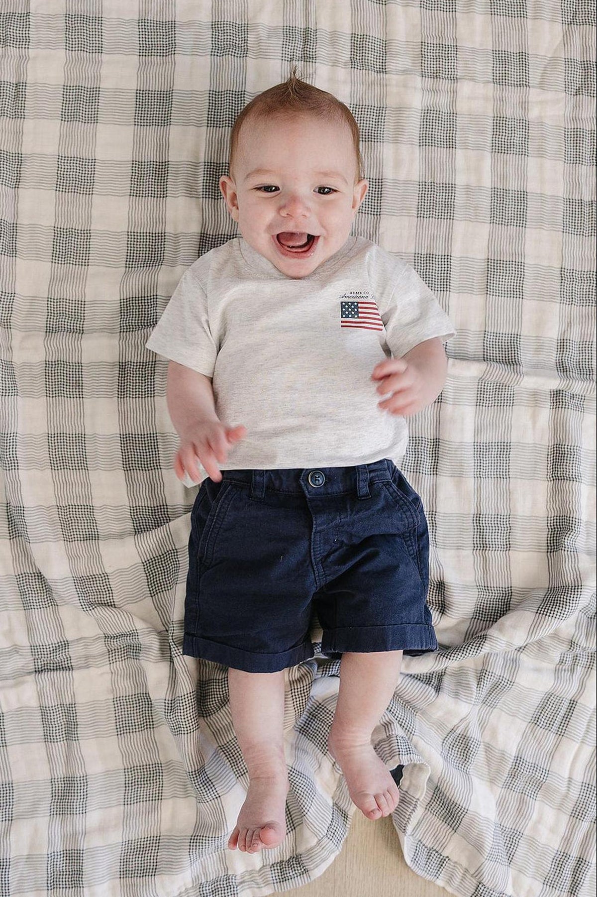 A smiling baby wears a gray T-shirt with an American flag graphic and navy shorts on a plaid blanket.