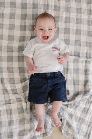 A smiling baby wears a gray T-shirt with an American flag graphic and navy shorts on a plaid blanket.