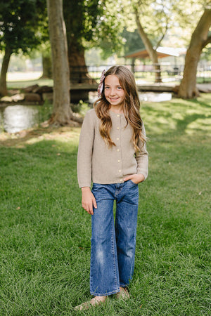A young girl stands on green grass near a pond, wearing a light brown knit cardigan with buttons and blue wide-leg jeans.