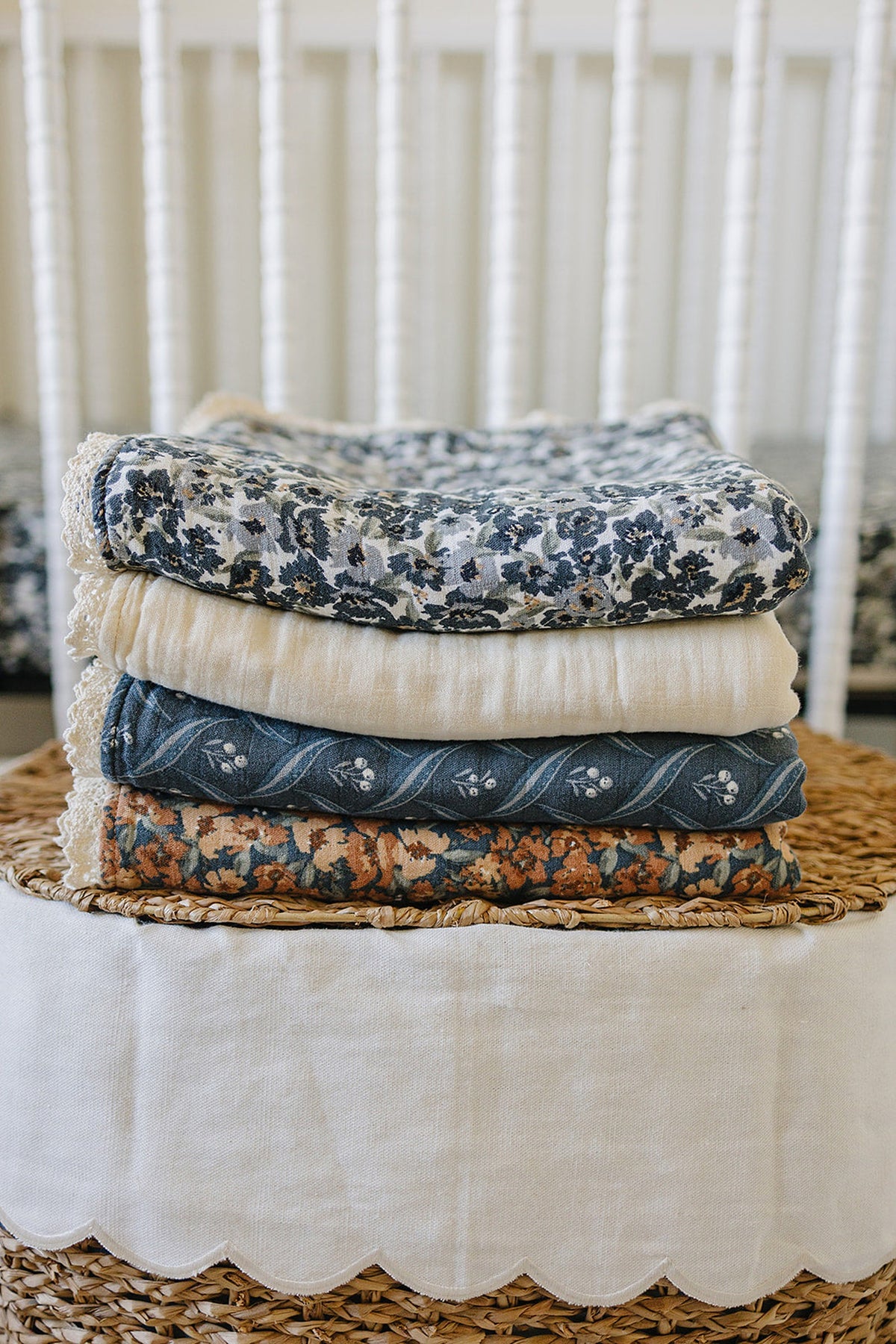 Stack of floral and plaid muslin quilts with lace edges on a wicker basket in a nursery setting.