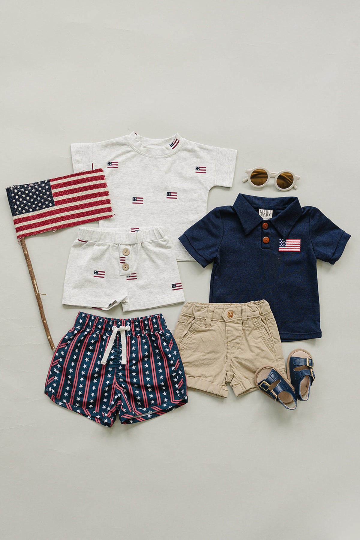 A flat lay displays various childrens clothing items, including a navy polo with a flag detail, a white shirt with flag patterns, striped shorts, khaki shorts, sunglasses.