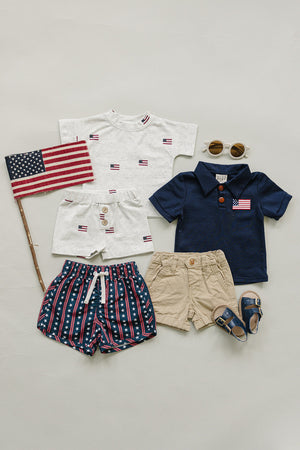 A flat lay displays various childrens clothing items, including a navy polo with a flag detail, a white shirt with flag patterns, striped shorts, khaki shorts, sunglasses.