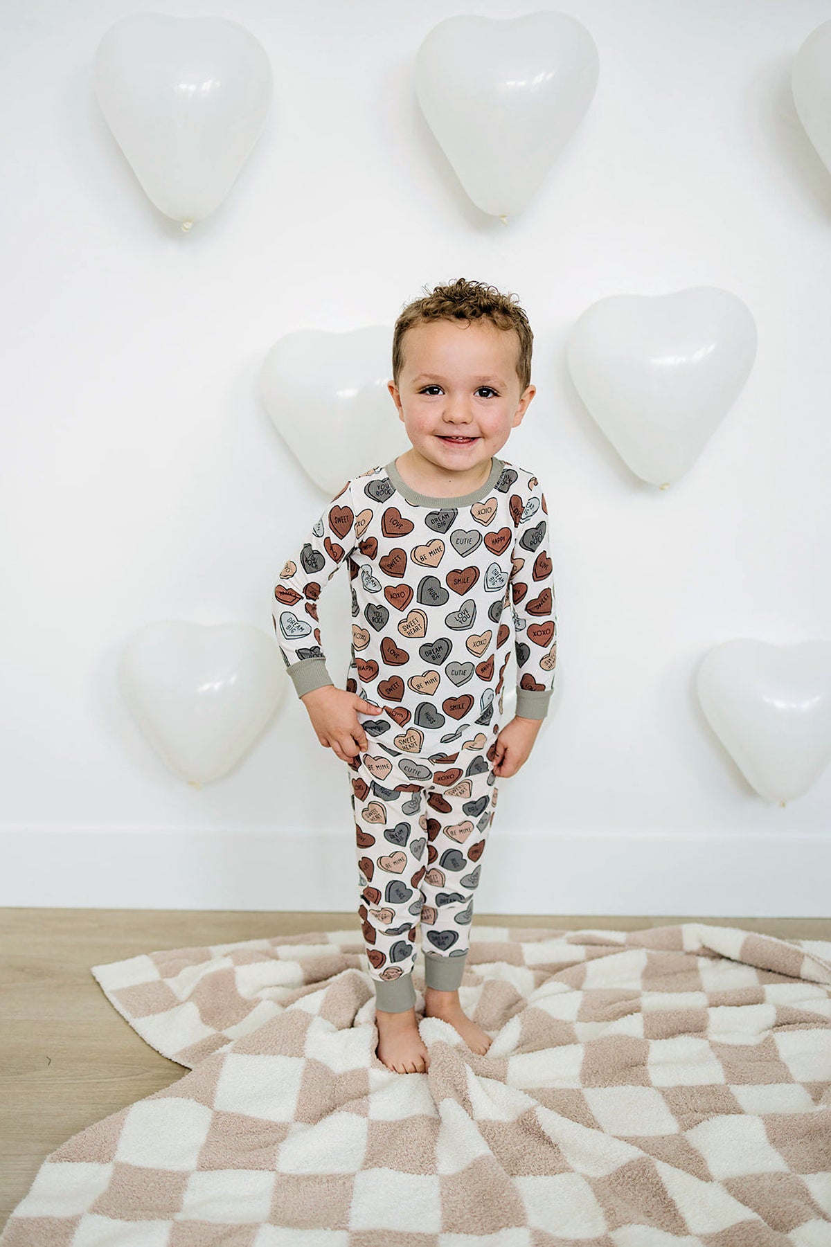 A smiling child wears a cozy pajama set featuring colorful heart prints, standing on a soft, checkered blanket, with heart-shaped.