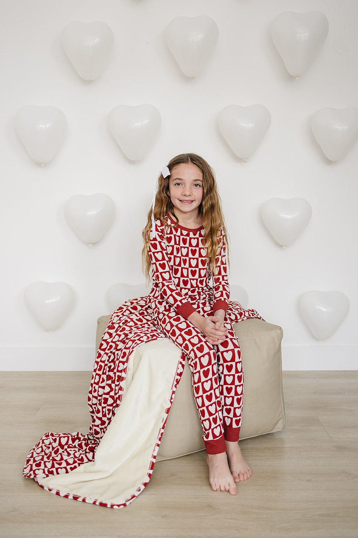 A child sits on a beige pouf, wearing a red checkered heart pajama set with a cozy blanket draped beside.