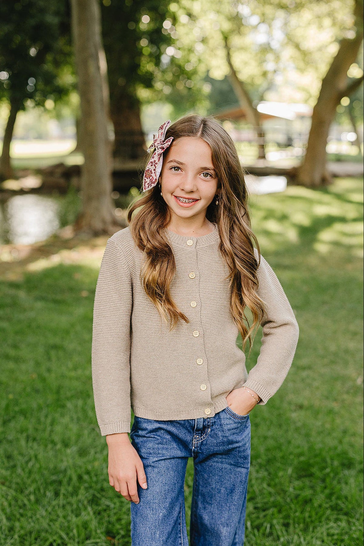 A young girl stands in a sunny park, wearing a beige knit cardigan with buttons, paired with blue jeans.