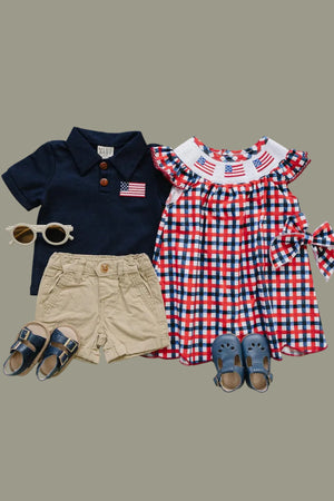 A navy polo shirt with an American flag patch, a red and blue plaid dress with a smocked collar, tan shorts, sunglasses.