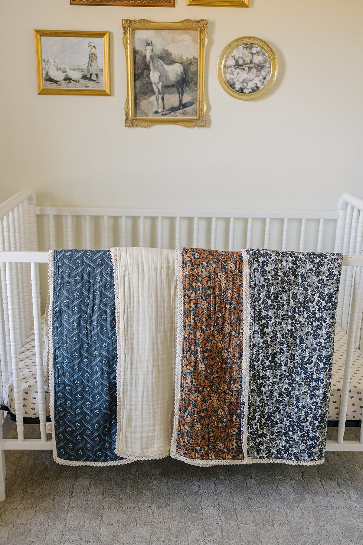 Four patterned muslin quilts with lace edges draped over a white crib, with framed art on the wall.