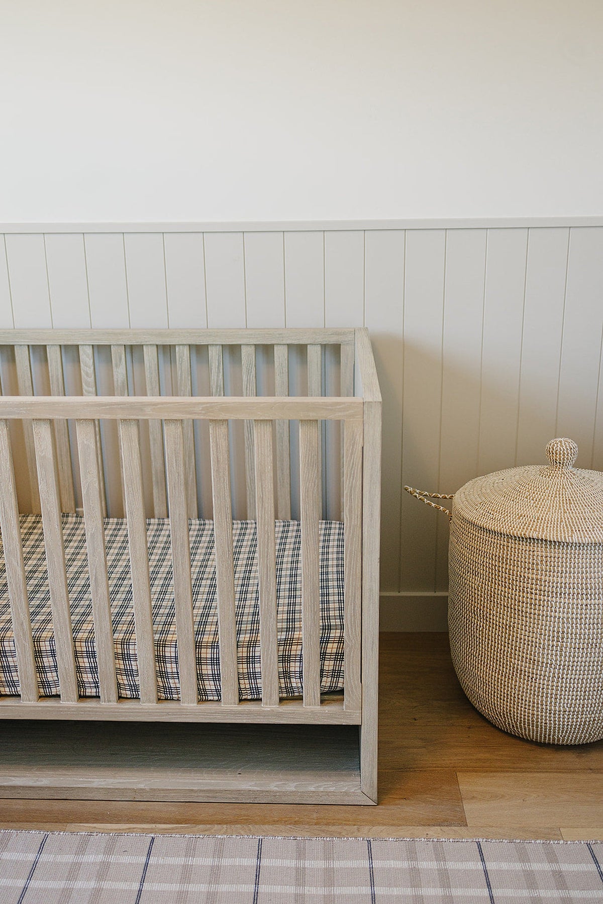 A light wooden crib with a plaid muslin sheet, next to a woven basket on a wooden floor.