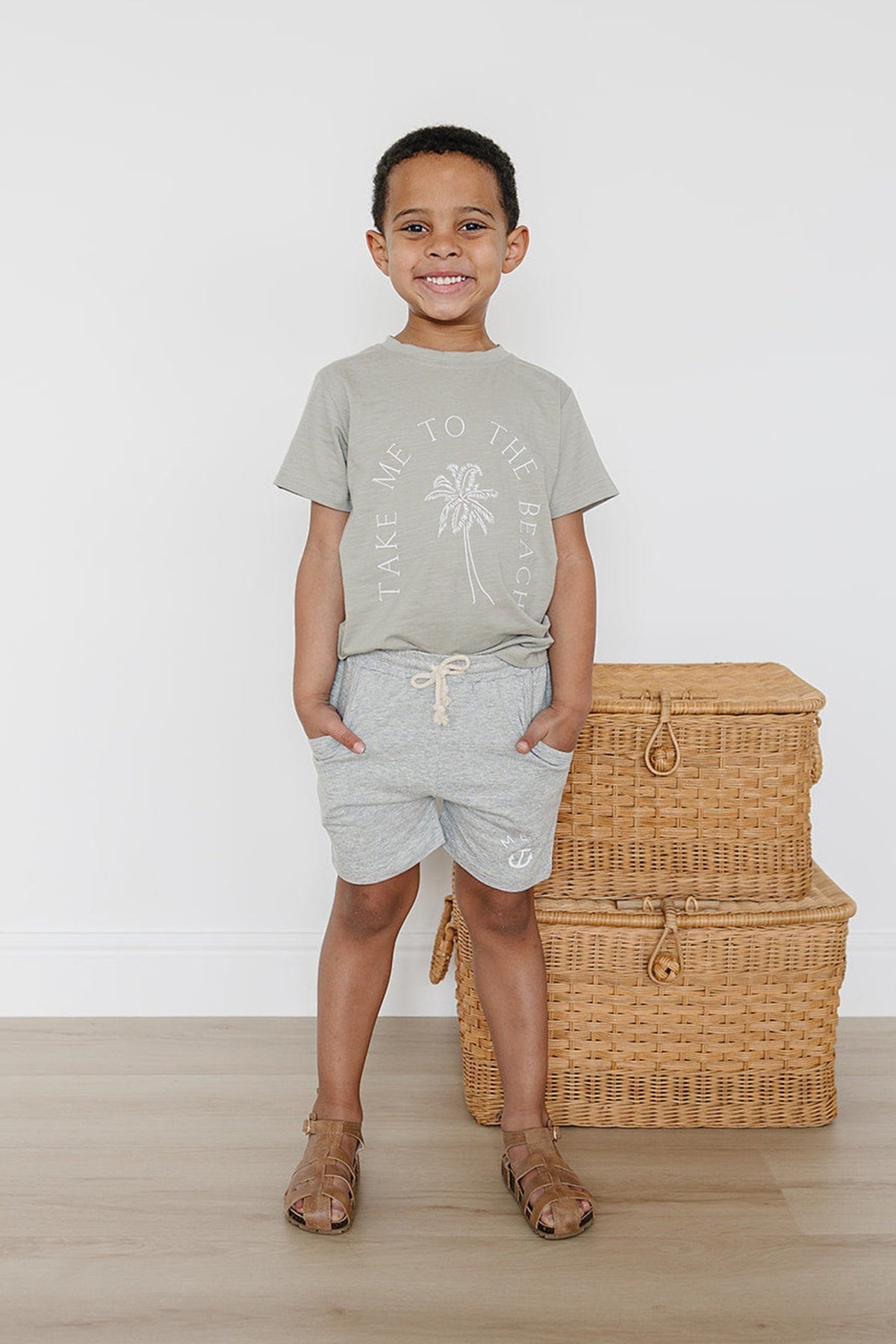 A child wearing heather grey cotton shorts and a beach-themed T-shirt stands beside wicker baskets, smiling brightly.