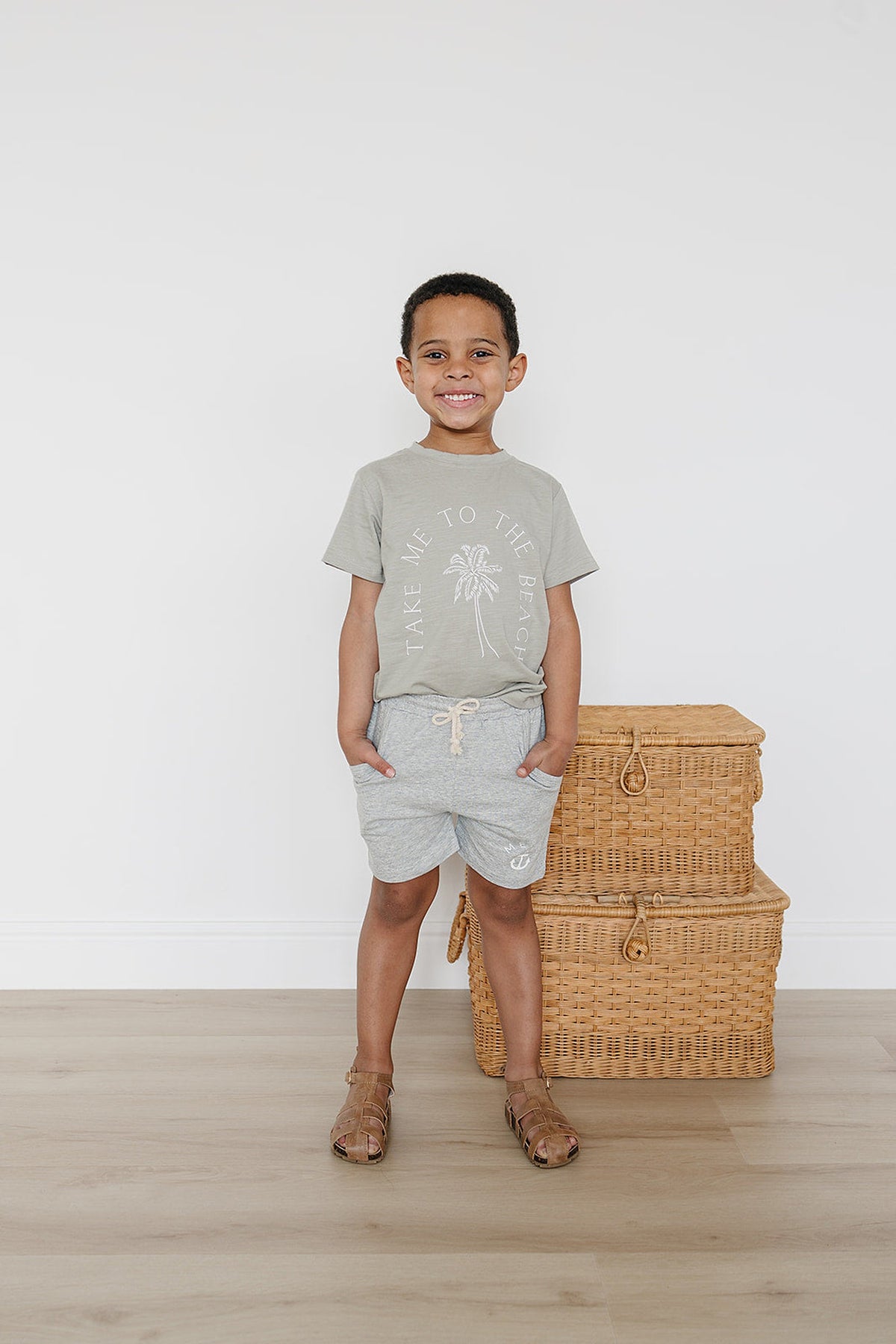 A smiling child wears a gray Take Me To The Beach t-shirt, standing beside wicker baskets on a wooden floor.