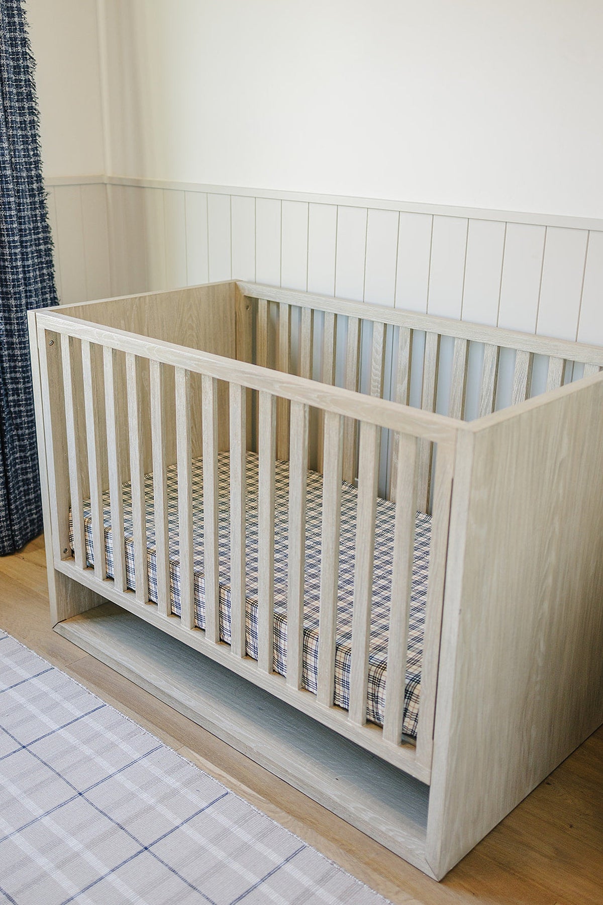 Light wood crib with vintage plaid muslin sheet, set in a nursery with paneled white walls and plaid rug.