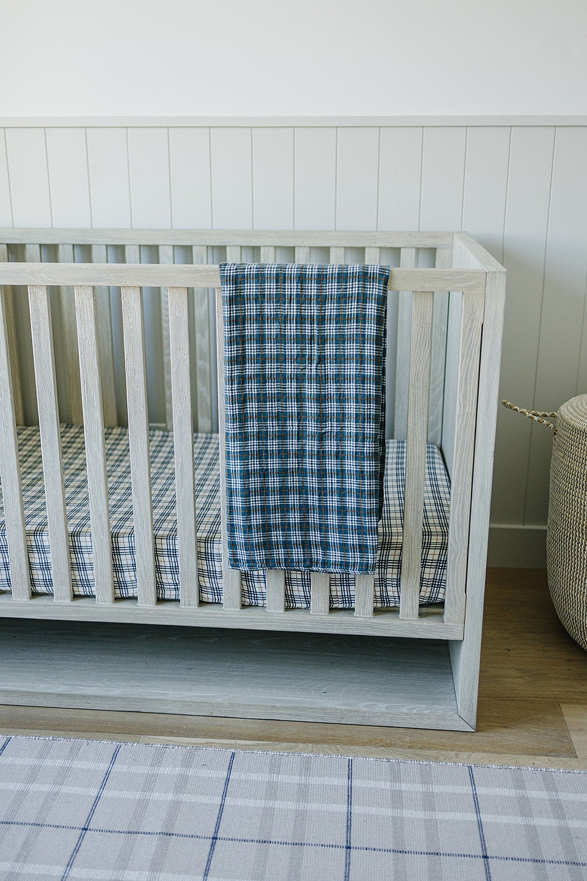A wooden crib with a navy plaid muslin quilt draped over the side, set in a nursery.