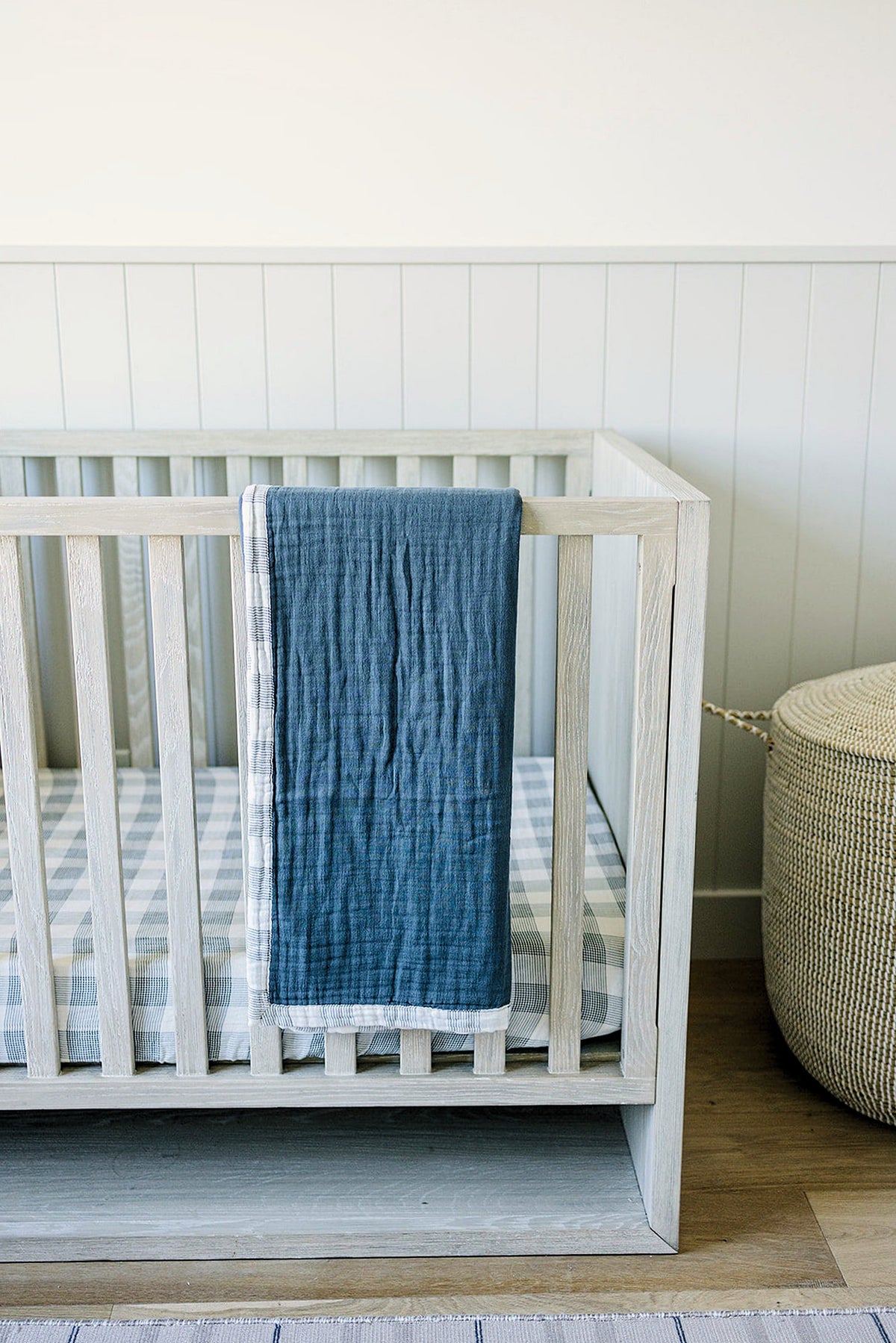 A light wooden crib with a navy and plaid muslin quilt draped over the side.
