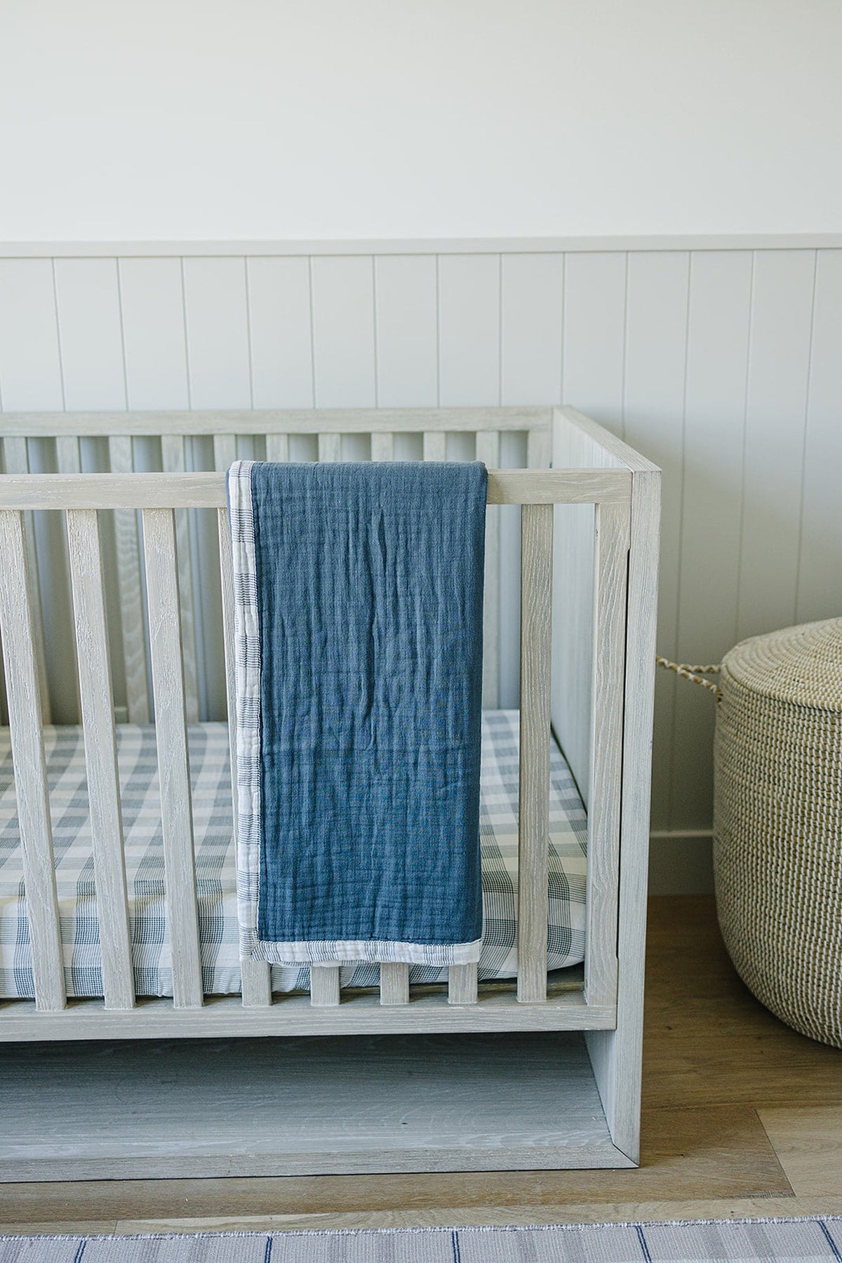 A gray muslin blanket hangs over a light wooden crib, which features a plaid patterned mattress.
