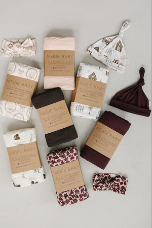 A variety of baby essentials from Mebie Baby, including swaddles and hats in soft colors and playful patterns, are neatly arranged with eco-friendly packaging on a neutral backdrop.