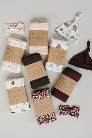 A flat lay arrangement of various baby essentials, including swaddles and hats in soft fabrics. The items feature neutral tones and whimsical patterns, showcasing Mebie Babys gentle, sustainable materials.
