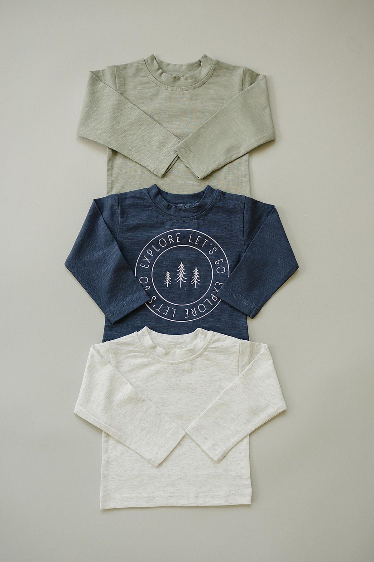 Three long-sleeve cotton shirts in sage, navy with graphic print, and cream are laid out on a neutral background.