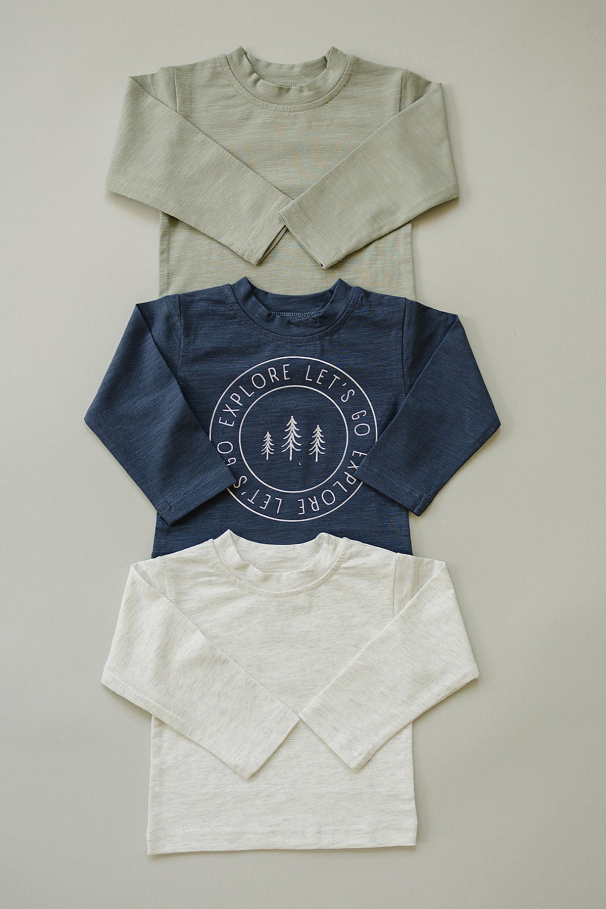 Three long sleeve shirts in green, navy with Lets Go Explore print, and cream, arranged on a flat surface.