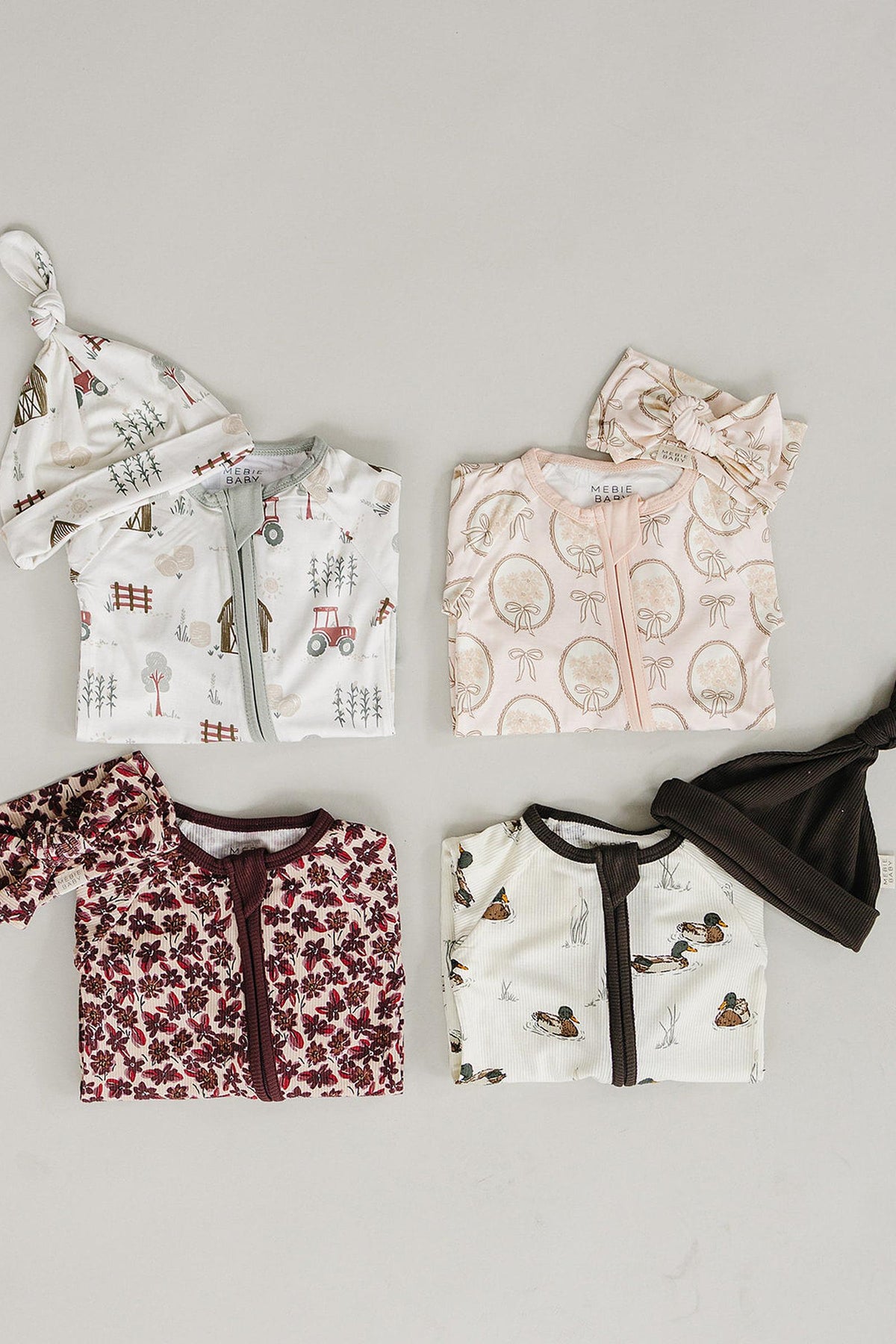 A collection of four baby outfits featuring various patterns, including floral, tractors, and woodland themes. Each outfit is designed with a convenient zipper and includes coordinating hats.
