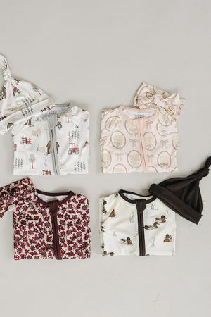 A flat lay displays four baby pajamas in soft bamboo fabric, featuring various whimsical prints, including tractors and mushrooms. Each pajama is paired with matching hats, emphasizing comfort and style.