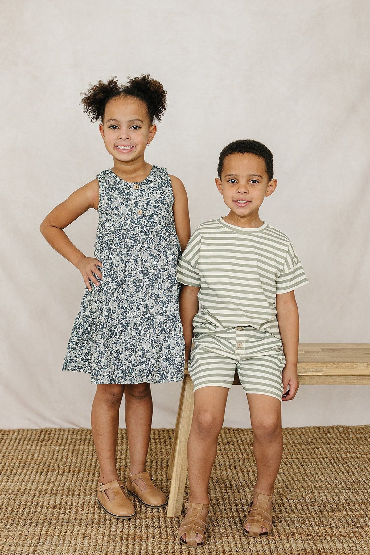 A girl in a floral tank dress and a boy in striped shorts stand on a wooden surface against a neutral backdrop, showcasing playful, stylish outfits for children.