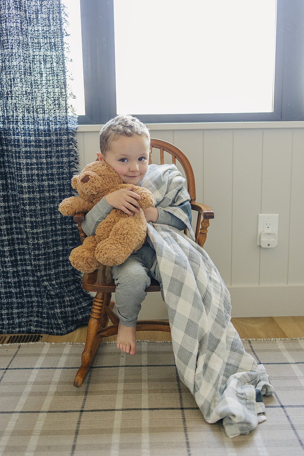 A child in gray pajamas sits on a wooden chair, holding a teddy bear and a plaid blanket.