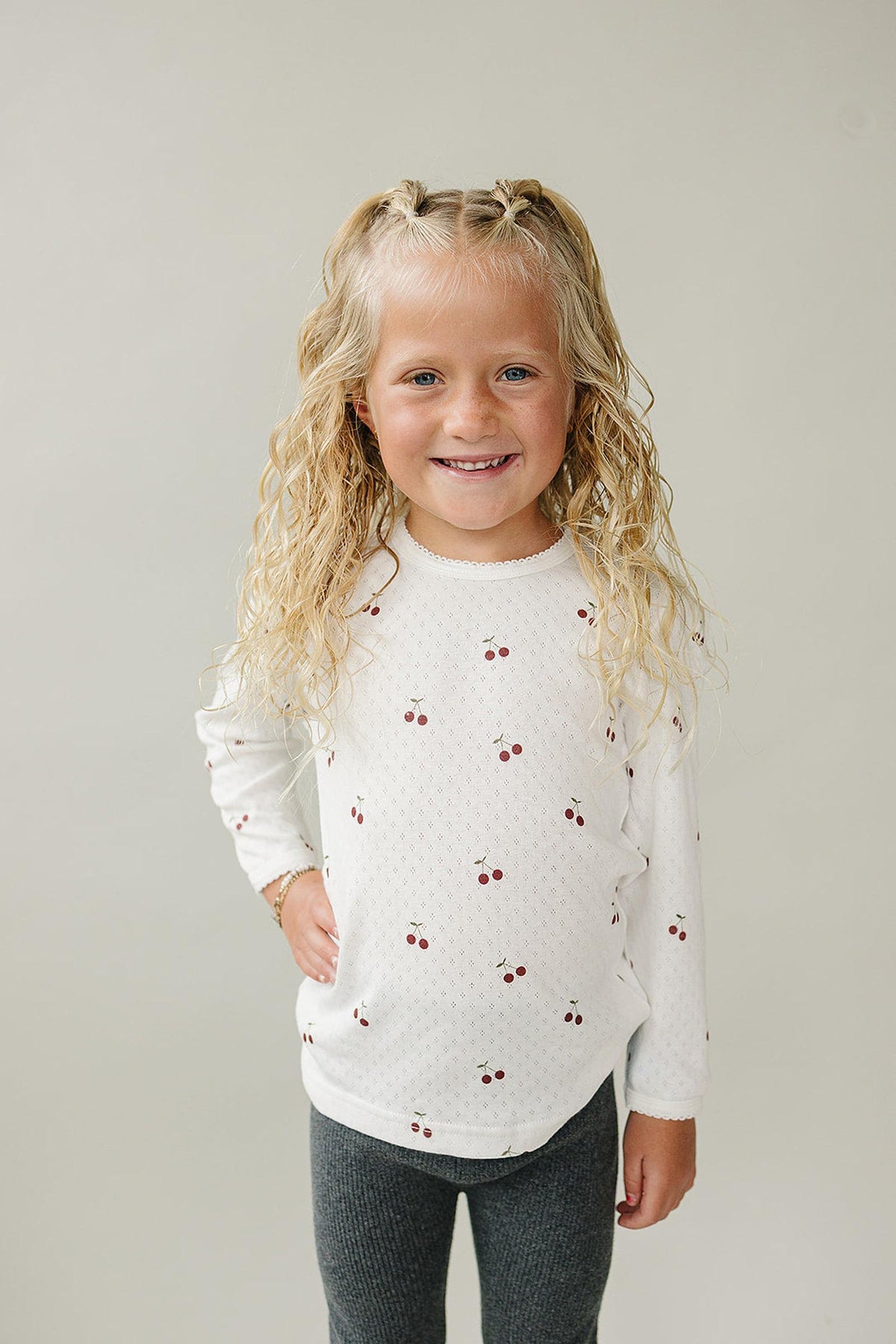 A smiling child with curly hair wears a white long sleeve shirt featuring cherry prints, paired with gray leggings.