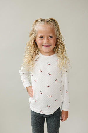 A smiling child with curly hair wears a white long sleeve shirt featuring cherry prints, paired with gray leggings.