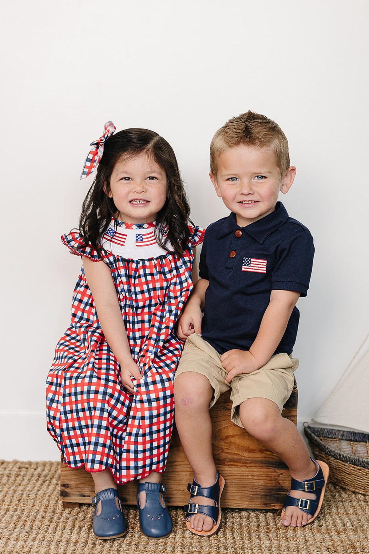 A girl in a red and blue gingham dress with an embroidered collar sits next to a boy in a navy polo shirt and khaki shorts.