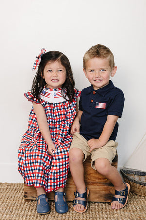 A girl in a red and blue gingham dress with an embroidered collar sits next to a boy in a navy polo shirt and khaki shorts.
