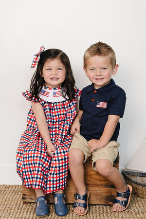 A girl in a red and blue plaid dress smiles beside a boy in a navy polo shirt with an American flag.