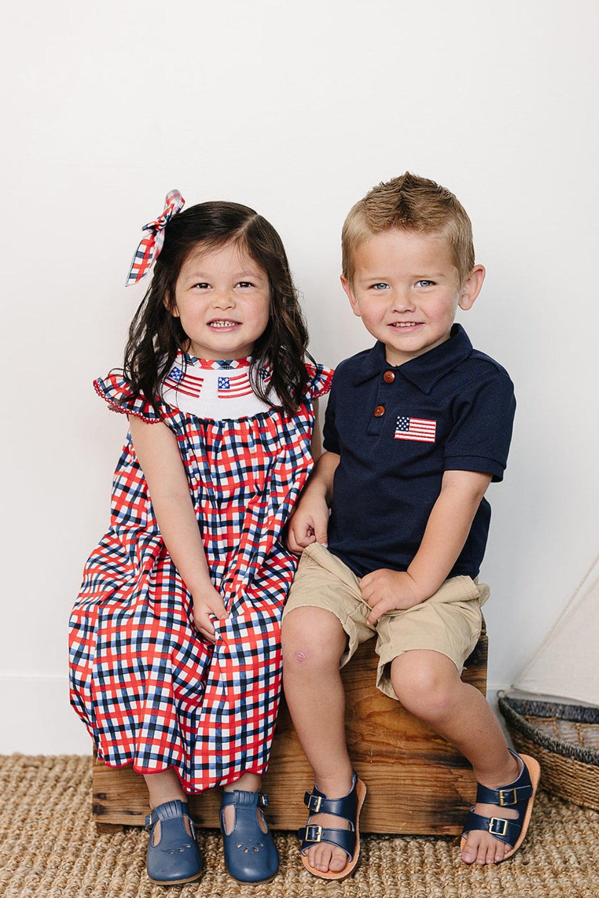 A girl in a red and blue checked dress and a boy in a navy polo sit on a wooden crate, smiling against a light background with nautical decor.