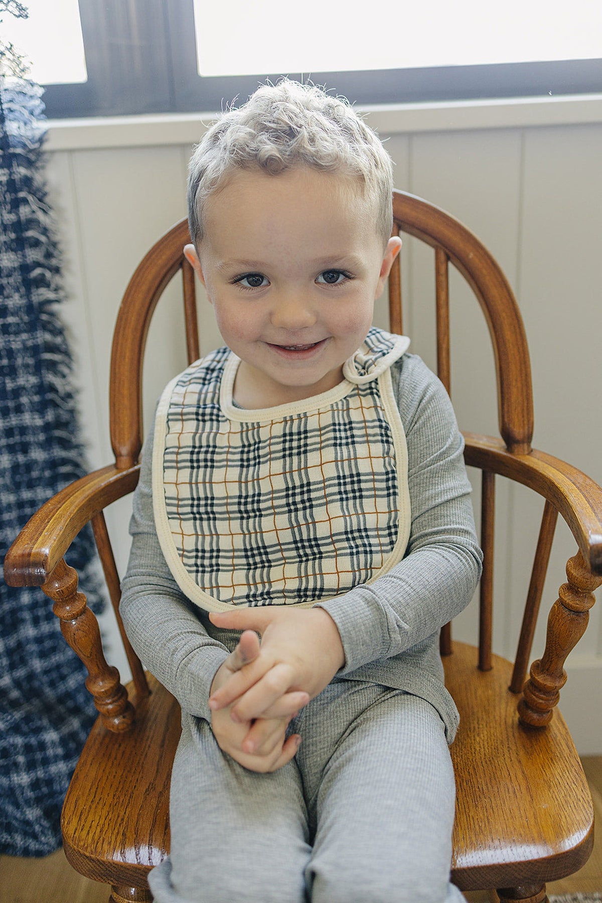 A child sits on a wooden chair wearing a plaid muslin bib and gray outfit, smiling warmly at the camera.