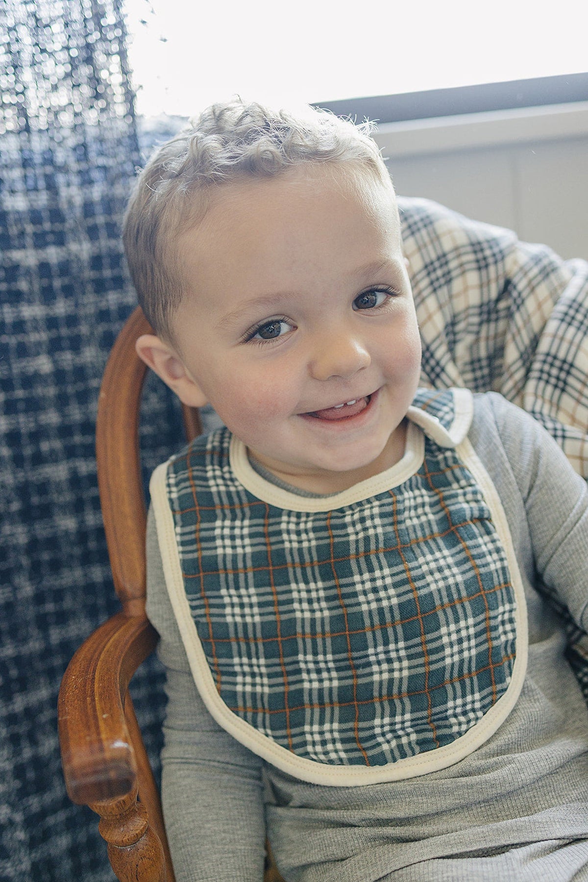 A smiling child wearing a navy plaid muslin bib sits in a wooden chair, with a plaid background.