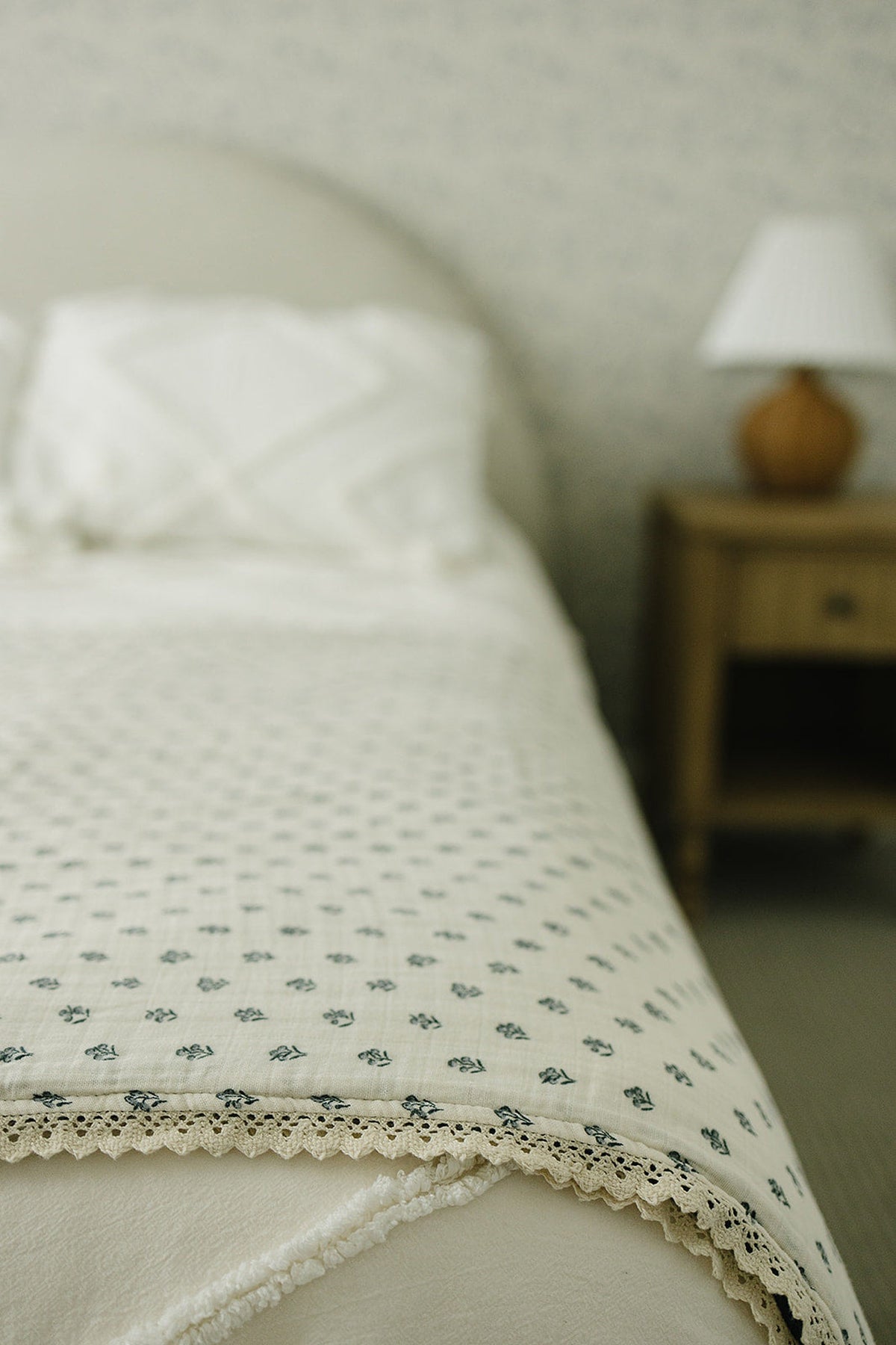 A cream quilt with a lace edge and navy berry pattern on a bed, near a wooden nightstand.