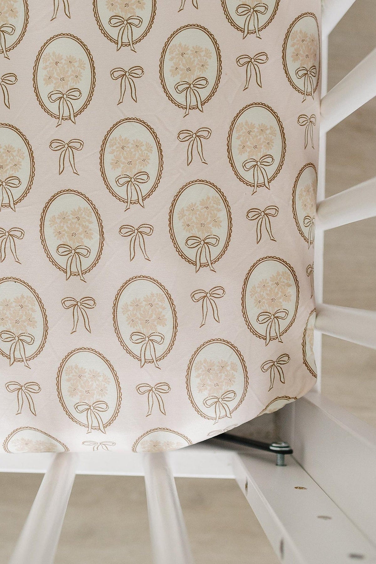 A vintage-style crib sheet features a delicate pattern of gold bows and floral motifs on a soft, pale pink background. It is designed to fit standard crib sizes.