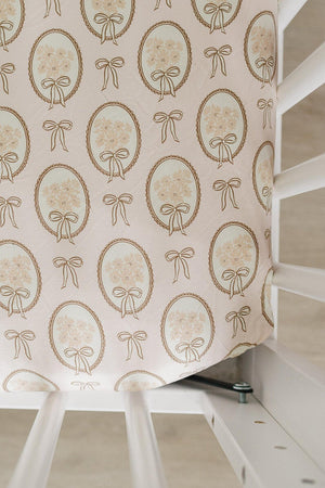 A vintage-style crib sheet features a delicate pattern of gold bows and floral motifs on a soft, pale pink background. It is designed to fit standard crib sizes.