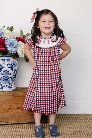 A smiling girl wears a red and blue gingham dress with a white smocked collar featuring embroidered American flags.
