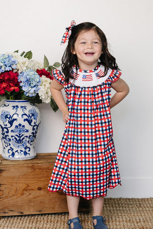 A smiling girl in a red and blue plaid dress stands against a neutral wall, wearing a matching plaid hair bow.