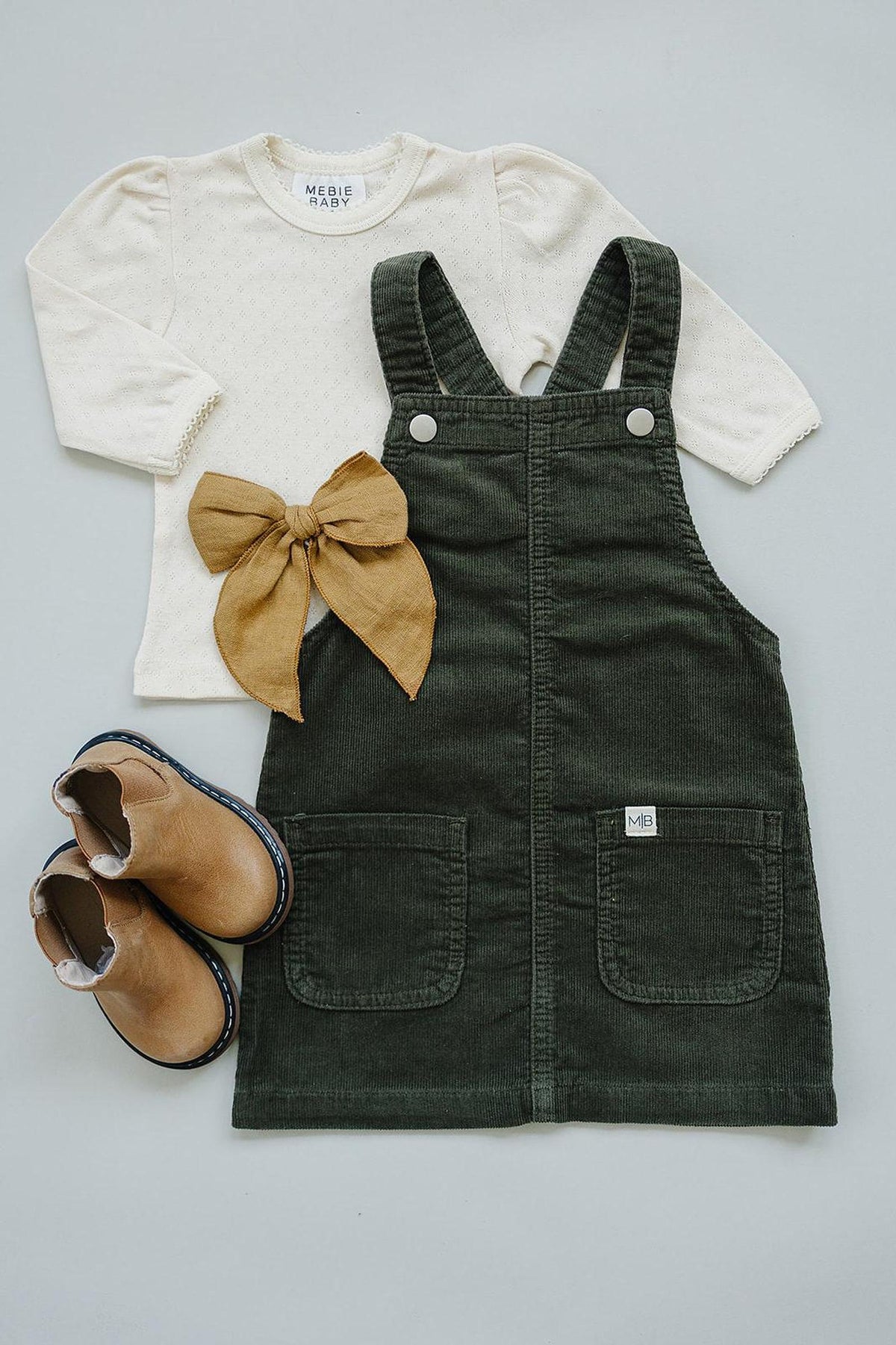 Cream long-sleeve top with a brown bow, green corduroy dress, and tan boots arranged on a flat surface.
