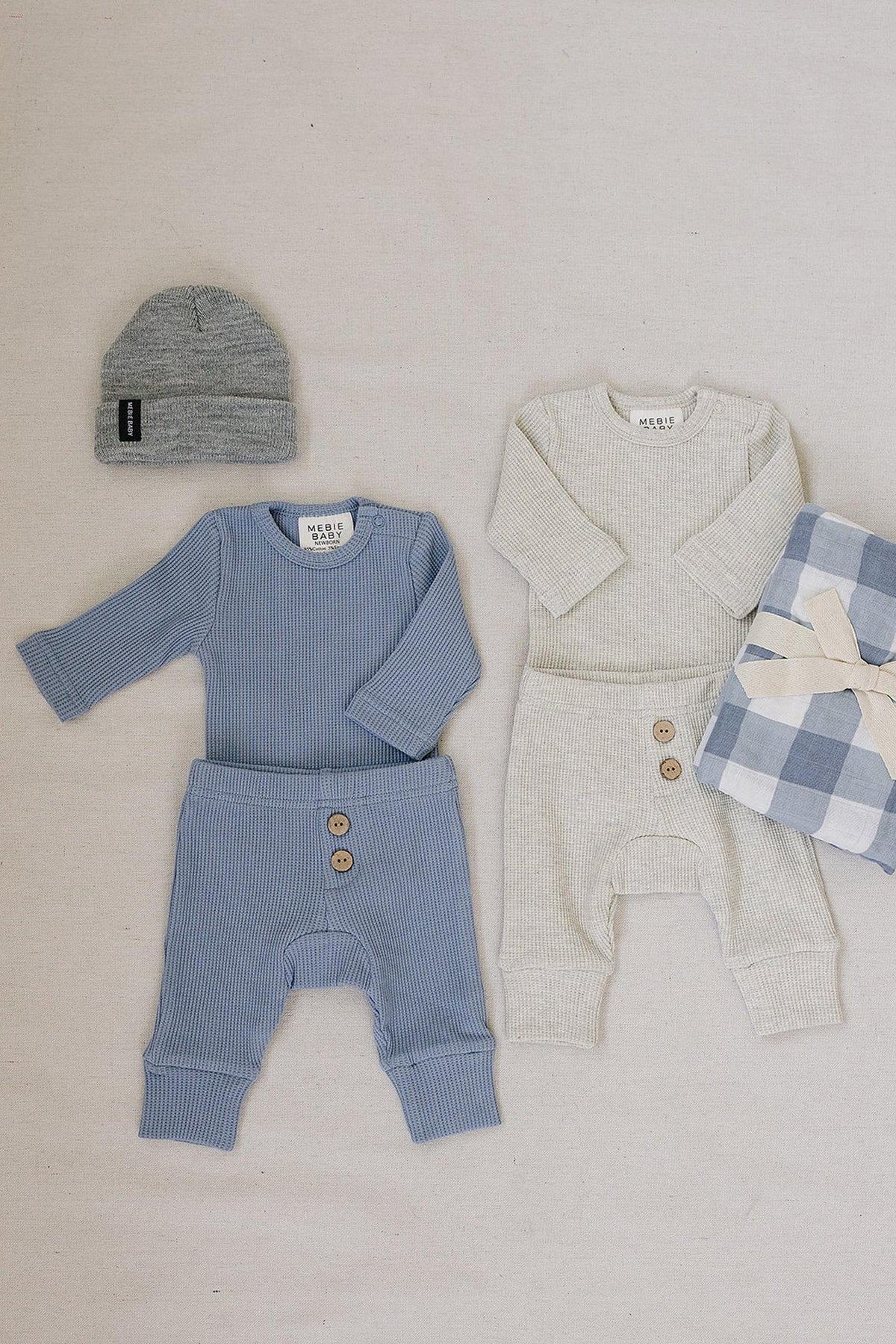A dusty blue waffle knit baby set is displayed alongside a light gray version, both featuring long sleeves and button details.