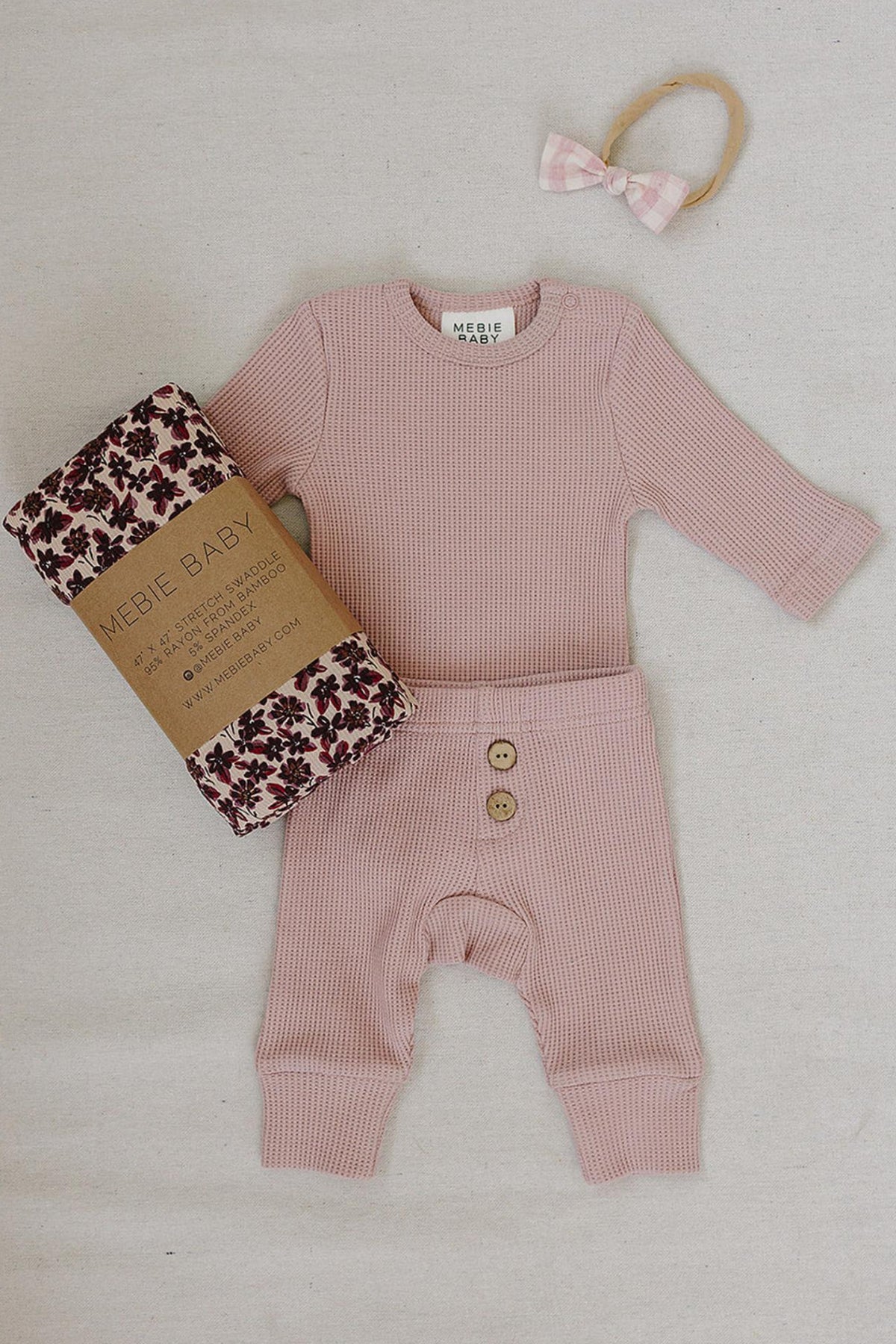 Dusty pink waffle knit cotton baby set featuring a long-sleeve bodysuit and relaxed fit pants, accompanied by a floral-patterned swaddle and a matching hair accessory. Perfect for comfort and style.