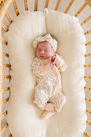 A peaceful newborn sleeps in a woven bassinet, wearing a soft, cream-colored zip-up onesie adorned with a whimsical mushroom print and a matching head wrap, showcasing gentle, organic materials.