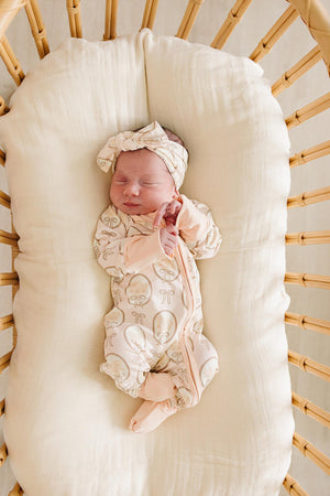 A sleeping baby in a bamboo zipper onesie with a matching headband lies peacefully in a cozy bassinet. The fabric features a soft, neutral color and playful mushroom print.