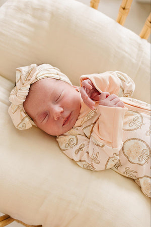 A peaceful newborn lies on a soft cream cushion, dressed in a vintage floral bamboo zipper pajama set with matching headband.