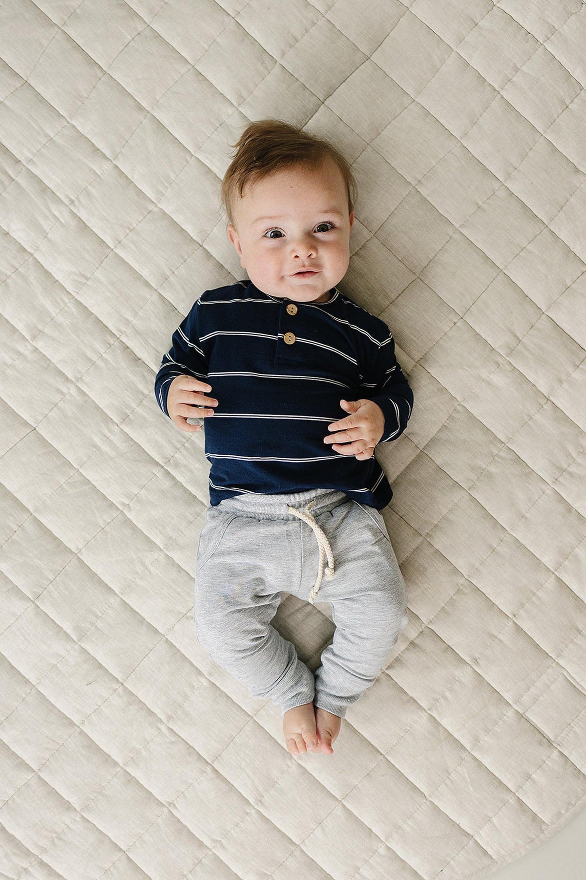 A baby lies on a textured, light-colored surface, wearing a navy long-sleeve shirt with white stripes and gray pants. The shirt features a two-button placket for added style.