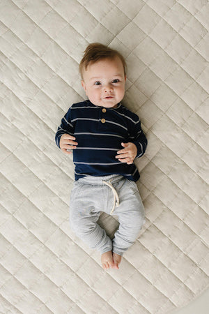 A baby lies on a textured, light-colored surface, wearing a navy long-sleeve shirt with white stripes and gray pants. The shirt features a two-button placket for added style.