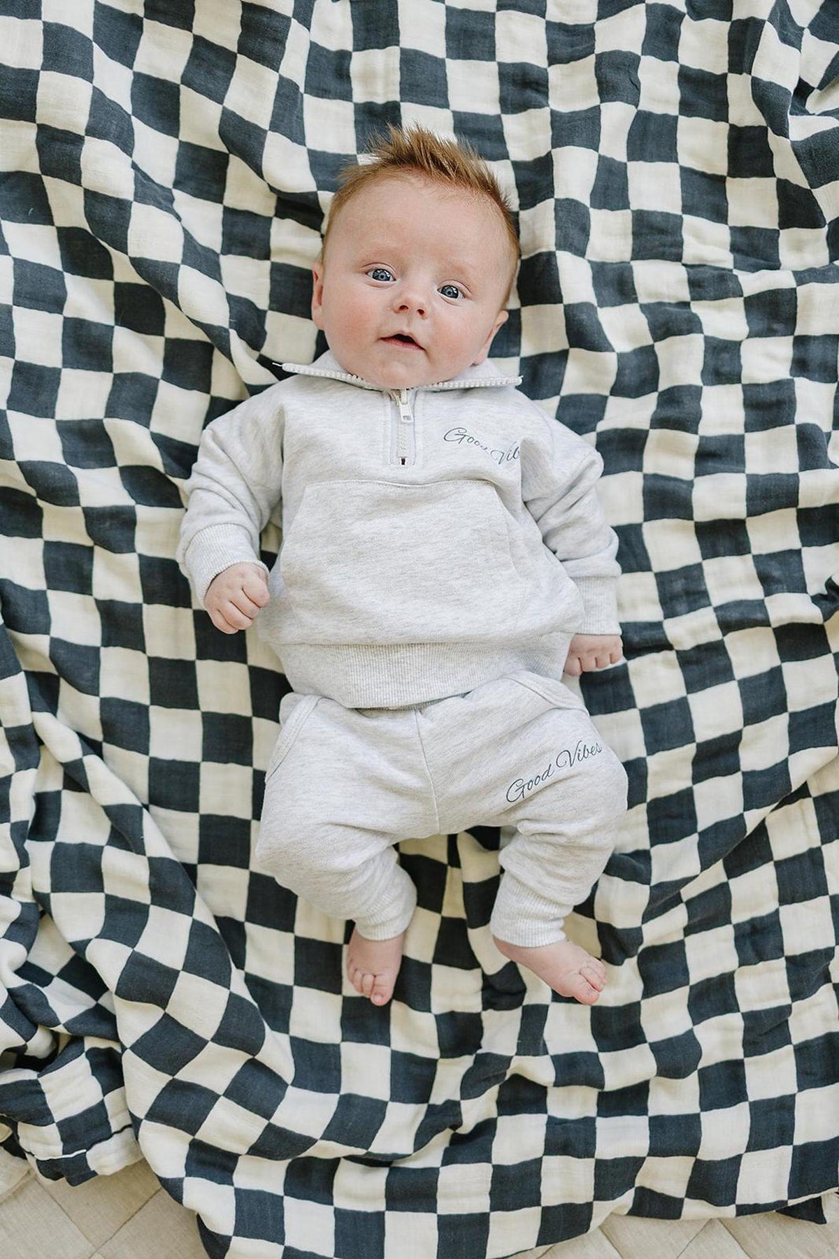 Baby wearing a grey quarter zip sweatshirt and pants with Good Vibes text, lying on a checkered blanket.