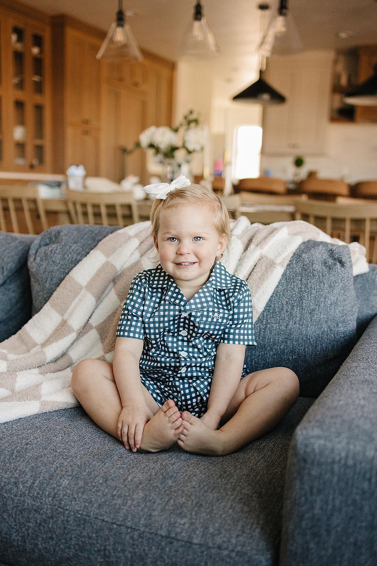 A smiling child in a navy gingham pajama set sits on a gray couch with a cozy blanket.