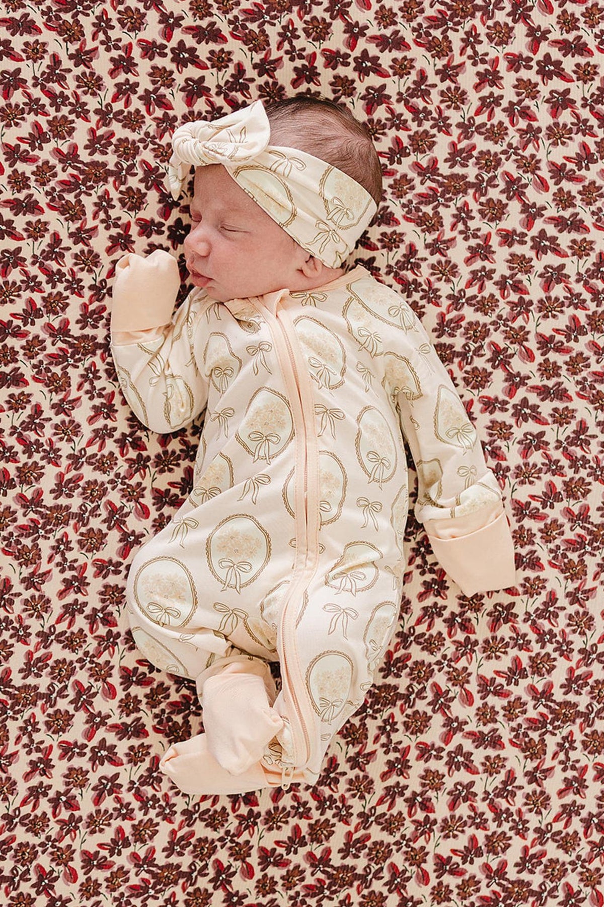 A newborn lies peacefully on a floral-patterned blanket, wearing a cream-colored bamboo zipper onesie adorned with whimsical mushroom prints and a matching headband. The soft fabric ensures comfort.